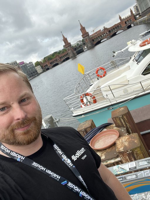 A bearded man wearing a 'Superteam' shirt and a 'Hacker House' lanyard takes a selfie in front of a harbor featuring a historic bridge with towers, likely the Oberbaumbrücke in Berlin.
