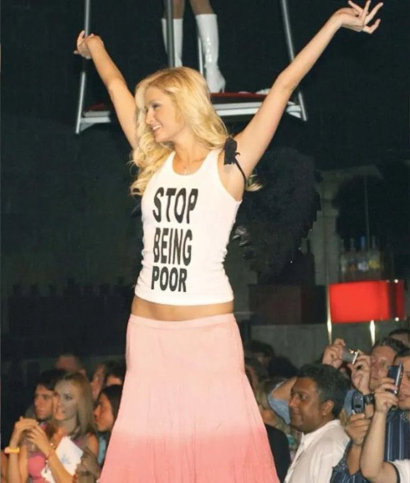 The image shows a woman standing on stage wearing a white tank top that says "stop being poor". She has her arms raised high, as if she's celebrating or expressing her opinion about the subject matter of the post. The crowd around her is watching attentively and some are taking pictures with their cell phones to capture the moment.