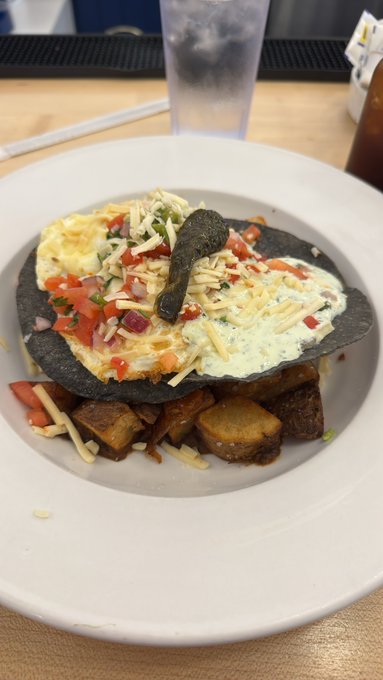 A plated breakfast featuring a black tortilla topped with eggs, cheese, and pico de gallo, served over roasted potatoes.