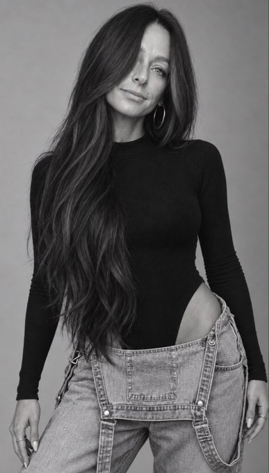 A black-and-white portrait shows a woman with long dark hair wearing a black long-sleeve bodysuit and open, unbuttoned denim overalls.