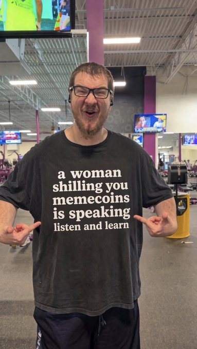 A bearded man in a gym wearing a t-shirt that reads "a woman shilling you memecoins is speaking listen and learn," pointing at the text with both hands.