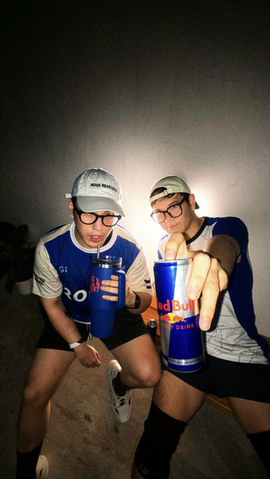 Two people pose at night wearing matching blue and white athletic jerseys, both drinking from Red Bull branded cups while striking playful poses.