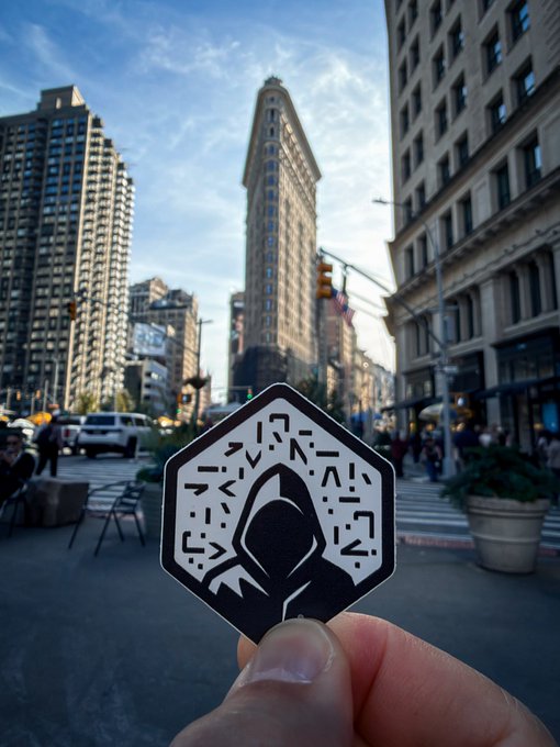 A hand holding a pentagonal sticker with an Assassin's Creed-style hooded logo and decorative patterns, set against a city street with the Flatiron Building in the background.