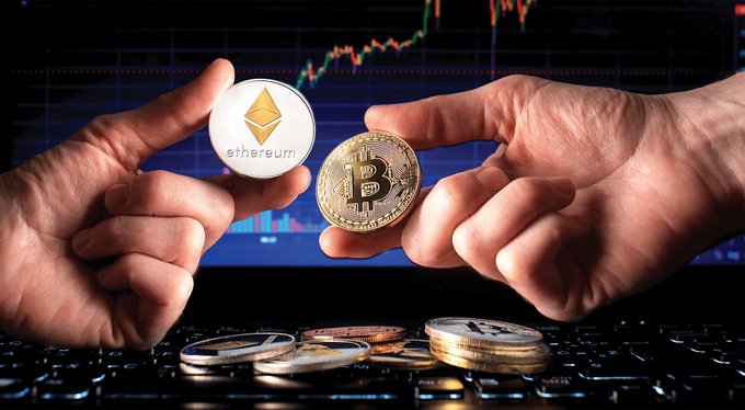 Two hands holding physical Ethereum and Bitcoin tokens in front of a laptop displaying a cryptocurrency price chart, with various crypto coins scattered on the keyboard.