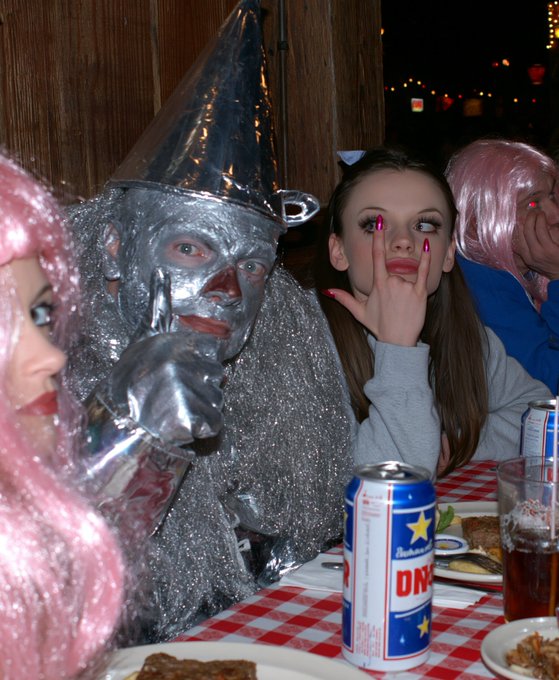 A person in a shiny silver Tin Man costume sits at a restaurant table next to a young woman making a hand gesture, with a person in a pink wig visible on the left edge. An 'AUSTIN' branded beer can and plates of food sit on the checkered tablecloth.