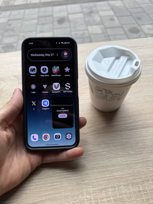 The image shows a person holding an iPhone with various apps displayed on its screen, including social media apps like Instagram and Twitter. The phone also has a coffee cup next to it, suggesting that the person might be enjoying their coffee while using their phone or taking a break from work or other activities.