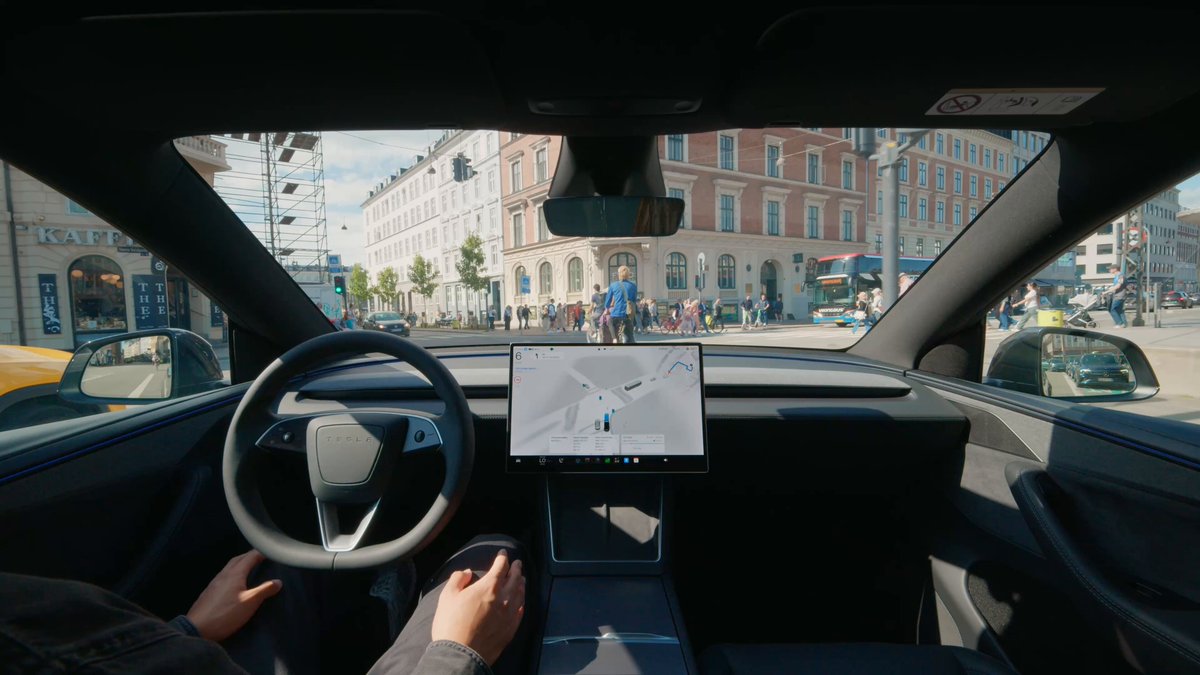 The image shows a person's hands driving a Tesla car with a large screen displaying a map of a city street. The car has a black interior, and there are other cars visible outside the vehicle. The caption reads "Just In: @Tesla has won approval for FSD (Supervised) in Denmark, the fourth European country to clear the system after the Netherlands, Lithuania and Estonia." This suggests that the person driving the Tesla is participating in an event or competition related to electric vehicles, possibly a race or demonstration of their car's capabilities.