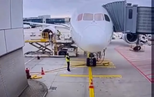 The image shows a large white airplane parked at an airport, with its nose pointed down towards the runway. A man wearing a yellow vest stands next to the plane, possibly working as a ground crew member or maintenance worker. The scene also includes several other airplanes in various positions around the airport, indicating that it is a busy location for air travel.