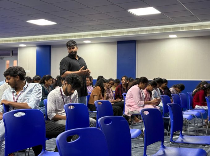 A bearded man stands and gestures while speaking to an audience seated in rows of blue chairs in what appears to be a conference or seminar setting. The image of a room full of attentive listeners doesn't directly relate to the post about the first NFT, "Quantum" by Kevin McCoy from 2014—though it may depict an event where such crypto/NFT topics are being discussed.