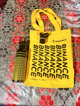 A yellow Binance-branded canvas tote bag with repeated 'BINANCE' text alongside a matching yellow water bottle, sitting on a patterned red and gray fabric surface.