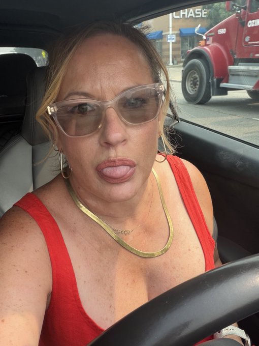 A woman in a red tank top and clear-framed sunglasses sticks her tongue out playfully from the driver's seat of a car, with a Chase bank sign and a red truck visible through the windshield—capturing her "good morning" selfie before stepping out to "touch grass" for the day.