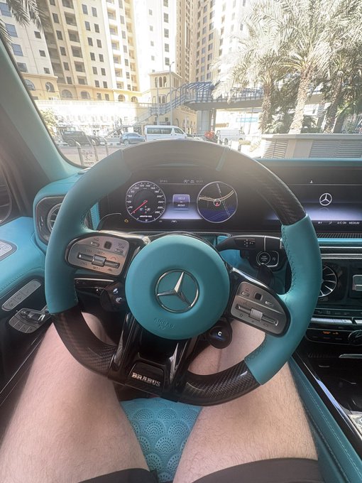 A first-person view from the driver's seat of a luxurious teal Brabus-tuned Mercedes, with the steering wheel, dashboard, and digital gauges clearly shown against a sunny urban backdrop. The post's "GM GM GM" likely refers to "Good Morning," greeting viewers with this stunning high-end interior.