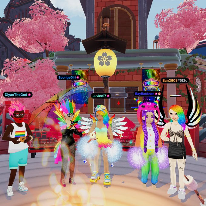 The image features five women dressed up in colorful costumes standing together in front of a building with pink flowers, possibly a temple or a palace. The group appears to be posing for a photo, showcasing their unique outfits and celebrating the LGBTQ+ pride event.