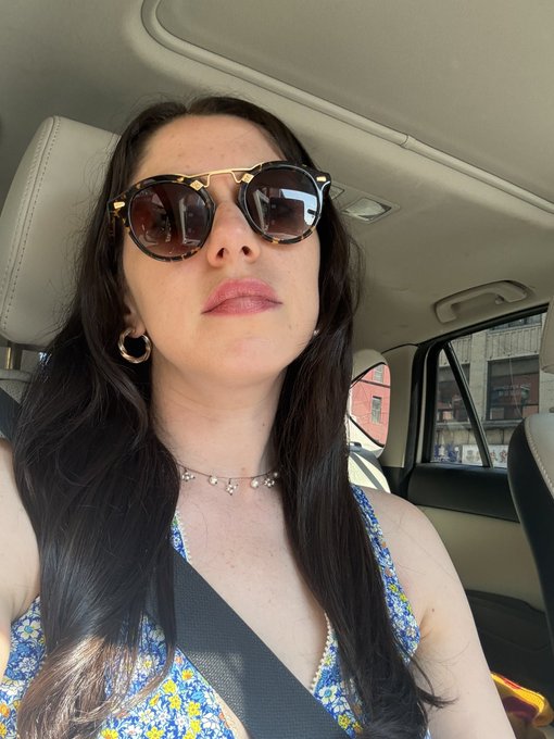 A woman takes a selfie in the car, wearing tortoiseshell sunglasses, gold hoop earrings, and a delicate charm necklace with a floral top, while they pause during a roadside stop to pick up fresh lychees.