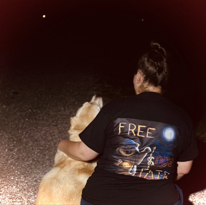 The image shows a woman sitting next to her dog, both of them wearing black t-shirts with messages written on them. One of the shirts has "Free Dogs" written on it, while the other shirt has "Dogs & Cats" written on it. The background is dark and out of focus, but there are some lights visible in the distance that could be streetlights or car headlights.