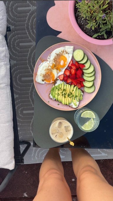 An overhead view of a healthy breakfast spread featuring fried eggs, avocado toast, strawberries, and cucumber on a pink plate, alongside an iced coffee drink.