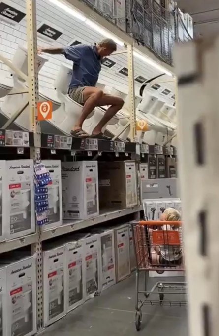 A man in a blue shirt and shorts sits perched on a top shelf in a hardware store aisle, surrounded by American Standard toilet boxes, with an orange shopping cart parked below.