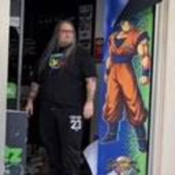 A large, bearded man in a black t-shirt and sweatpants with the number 23 stands next to a Dragon Ball Z poster, appearing to imitate the character's muscular pose.