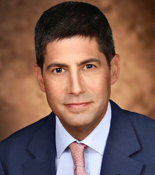 The image shows a man dressed in a suit and tie, wearing a serious expression as he looks directly at the camera with an unshakable gaze. The background of the picture appears to be brown, indicating that the photo may have been taken indoors or under artificial lighting conditions. There is no text present in the image, which seems to focus on the man's attire rather than any additional information about him.
