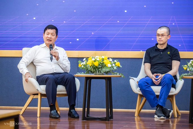 Two men seated on stage at a panel discussion, one in a white shirt holding a microphone and the other in a black polo shirt, with yellow flowers between them and a "VNEXPRESS" watermark.
