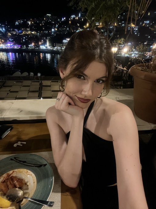 A woman with brown hair and dark lipstick rests her chin on her hand at an outdoor restaurant table at night, with a scenic illuminated coastal town visible in the background.