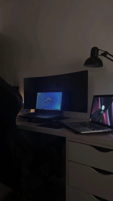 A dimly lit desk setup featuring a laptop, a curved monitor, a desktop lamp, and a gaming chair.