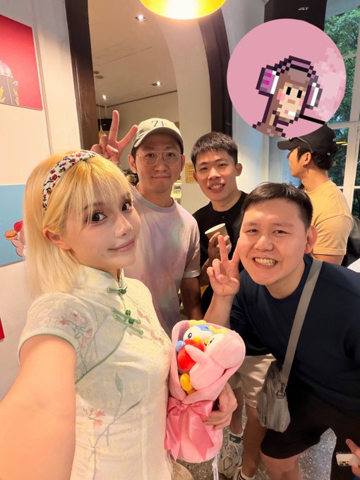 A group selfie featuring a blonde woman in traditional Chinese attire holding a pink plush toy, surrounded by three men making peace signs, with a pixelated cat avatar graphic overlaid.