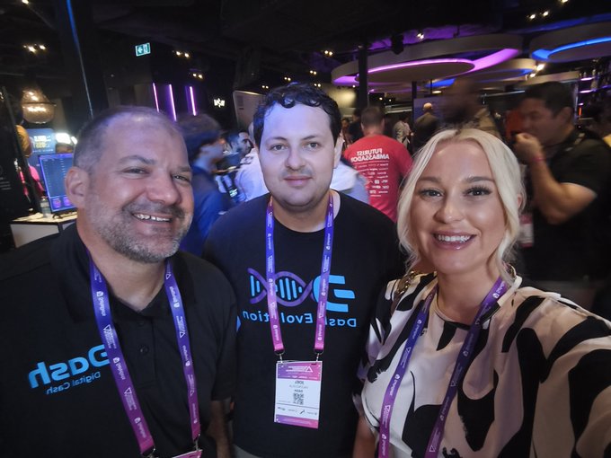 Three people pose together at what appears to be a conference; one man wears a black shirt with "DASH Evolution" and a "Digital Cash" logo, and all wear lanyards with conference badges.