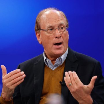 An older man with glasses wearing a dark suit jacket over a yellow sweater and striped shirt speaks animatedly with hand gestures against a blue background, appearing to be at a conference or interview.