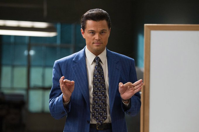 Leonardo DiCaprio, portraying Jordan Belfort in "The Wolf of Wall Street," stands in a pinstripe suit with both hands gesturing "OK" signs, conveying confident salesmanship. The image humorously illustrates the post's satirical take on shilling memecoins through a new L1 blockchain, embodying the sleazy, hyper-confident energy of Belfort's infamous "sell me this pen" pitch — applied to crypto rather than fountain pens.