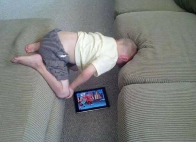 A baby in a cream shirt and gray pants lies face-down on a beige couch, resting their head near a tablet playing a video.