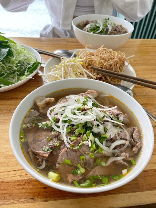 A traditional Vietnamese beef pho served in a white bowl, topped with sliced onions, green onions, and beef, accompanied by a side plate of bean sprouts, herbs, lime, and crispy noodles on a wooden table.