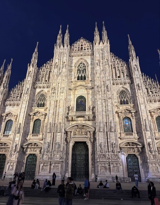 The illuminated facade of Milan Cathedral (Duomo di Milano) at night, featuring its ornate Gothic architecture with spires and statues, with people gathered in the plaza below.