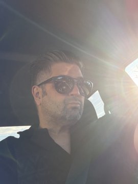 A man with short hair and sunglasses sitting in a car, photographed with bright sunlight streaming through the window creating lens flare.