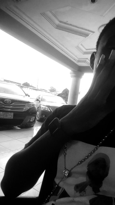 Black and white photo showing a person with a watch and chain jewelry, hand near face, with cars (Toyota visible) parked in the background.