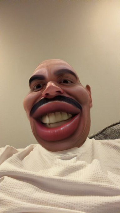 The caption could be: "An exaggerated, humorous filter distorts a man's face, enlarging his smile and lips, capturing the playful tone of going live while teasing a friend's car crash."

The image shows a man whose face has been comically warped by a filter, stretching his smile and features into an over-the-top grin. This matches the post's lighthearted, mocking vibe about streaming live alongside a friend's car mishap, using the absurd visual to amplify the joke.