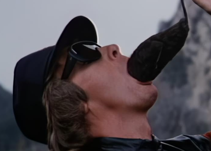 A man wearing a black cap and sunglasses with his head tilted back, appearing to drink or pour something from a black pouch or bag.