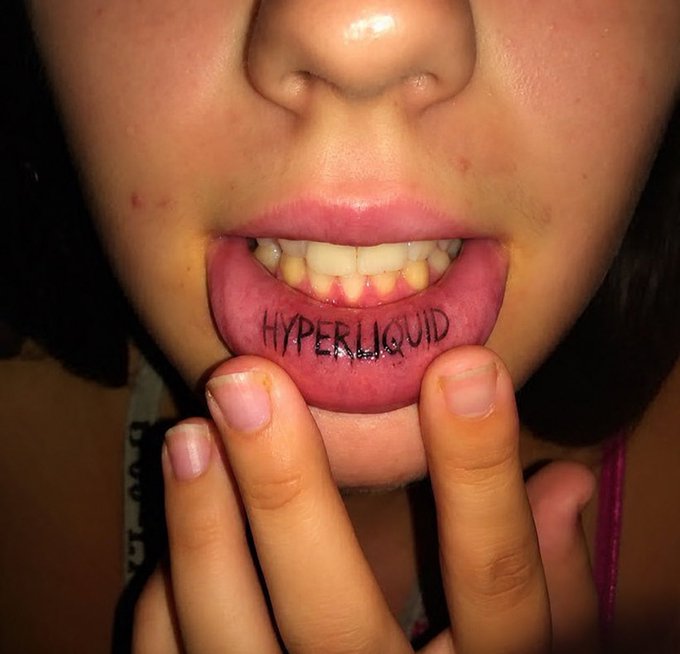 A close-up photograph of a person's mouth with "HYPERLIQUID" written in silver marker on their lower lip, with fingers pulling the lip down to reveal their teeth.