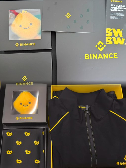 Binance branded merchandise collection including gray boxes with the Binance logo, a plush yellow mascot toy, and a black jacket with yellow Binance accents, tagged as "9YA GLOBAL AMBASSADOR CAMPAIGN".