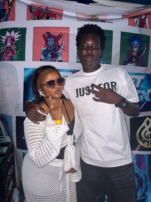 A young man and woman pose together at an anime convention booth featuring Yu-Gi-Oh! character artwork; the woman wears a white crocheted outfit and sunglasses, while the man wears a white 'JUST FOR' t-shirt.