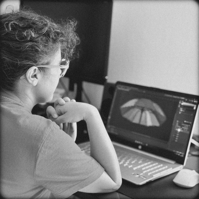 Black and white photo of a person with curly hair and glasses sitting at a desk, looking at 3D modeling software displayed on a laptop screen.