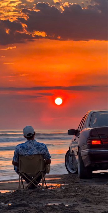 A scenic sunset view at a beach, featuring a man sitting in a folding chair facing the ocean, with a car parked next to him on the sand.