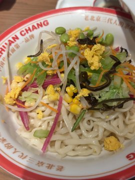 A bowl of "Huan Chang" (环长) fried sauce noodles topped with bean sprouts, corn, egg, cucumber, carrots, and seaweed sits in a branded bowl featuring Chinese characters like "京北三代行人" along the rim, matching the post's reference to Beijing Fangzhuan Factory's tasty fried sauce noodles that required waiting in line.