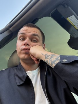 A selfie-style photo of a man with dark hair sitting in a car, wearing a black jacket over a white shirt, with his hand resting near his chin, displaying visible arm tattoos.