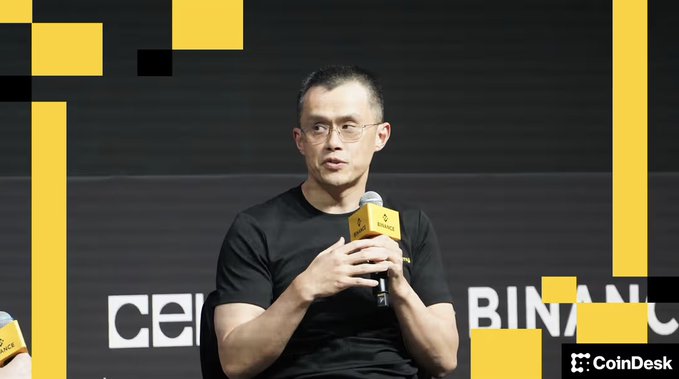 A man wearing glasses and a black t-shirt speaks into a Binance-branded microphone on stage at CoinDesk's Consensus event, with "cel" and "BINAN" visible on the backdrop behind him. The image accompanies the quote from CZ, founder of Binance, advocating for Bitcoin as protection against inflation over AI.
