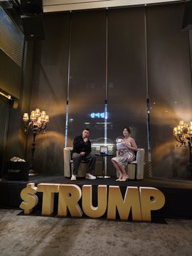 Two people sit on chairs on a stage behind large golden letters spelling '$TRUMP' in an elegant room.