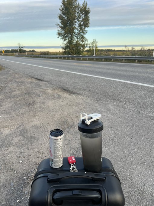 A black suitcase on a road shoulder serving as an impromptu table, holding a Diet Coke can, a water bottle, and car keys, with a rural highway and sunset in the background.