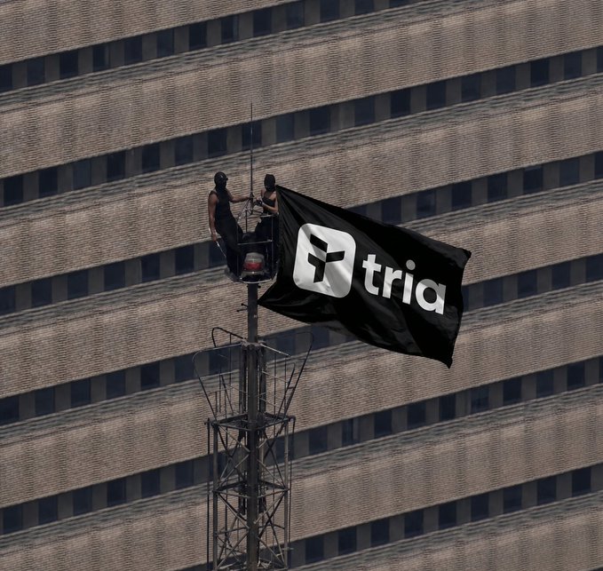 Two workers on a crane platform unfurling a large black flag with the FTX logo (now reading 'tria' due to defacement) against a building backdrop.