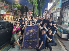 A large group of people posing together at night on a street, holding a blue banner with Thai or similar script and a crest.