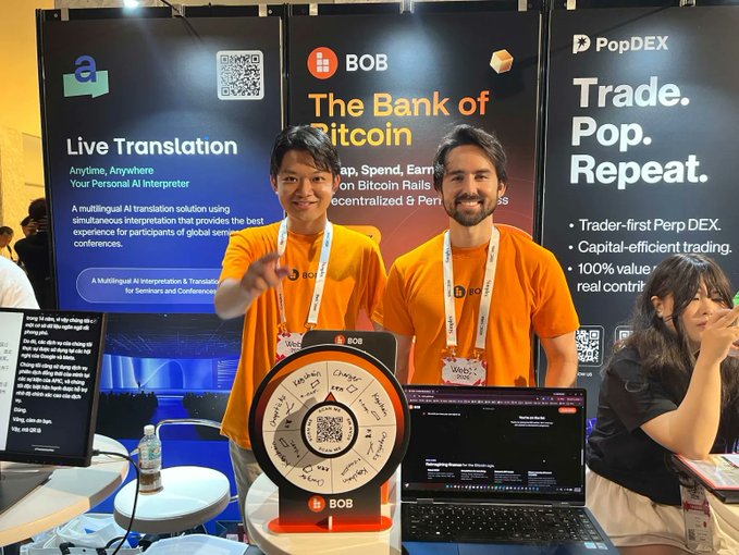 Two representatives in orange shirts stand at the BOB "Bank of Bitcoin" booth at WebX Asia in Tokyo, with a spinning prize wheel and signage for "Live Translation" and "PopDEX" visible in the background. The image captures the team's setup at Booth S-2 as promoted in the post, featuring interactive elements like the merch wheel to draw attendees.
